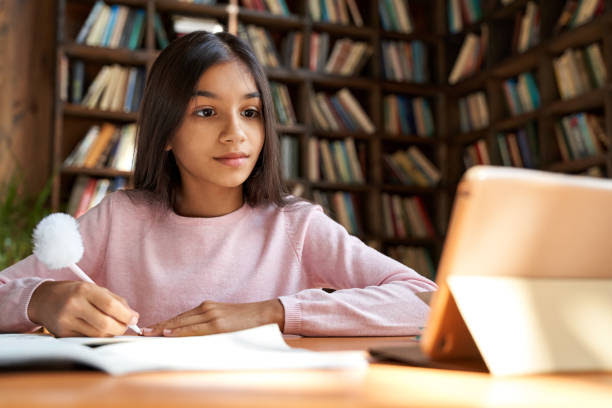 Indian latin girl school pupil distance learning online at remote virtual class with teacher by video conference call, watching webinar, meeting lesson on digital tablet at home, in classroom.